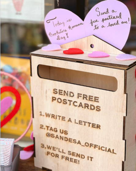 A wooden box with engraved text that reads: "Send free postcards. 1. Write a letter. 2. Tag us. 3. We'll sed it for free!